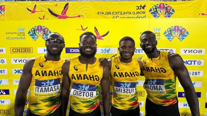 Four athletes pose before a yellow background, 他们穿着黄色的加纳球衣，手挽着手微笑着.